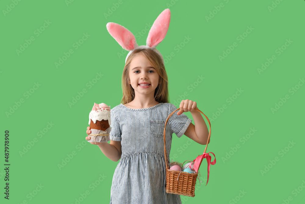 Little girl with tasty Easter cake and eggs in basket on green background