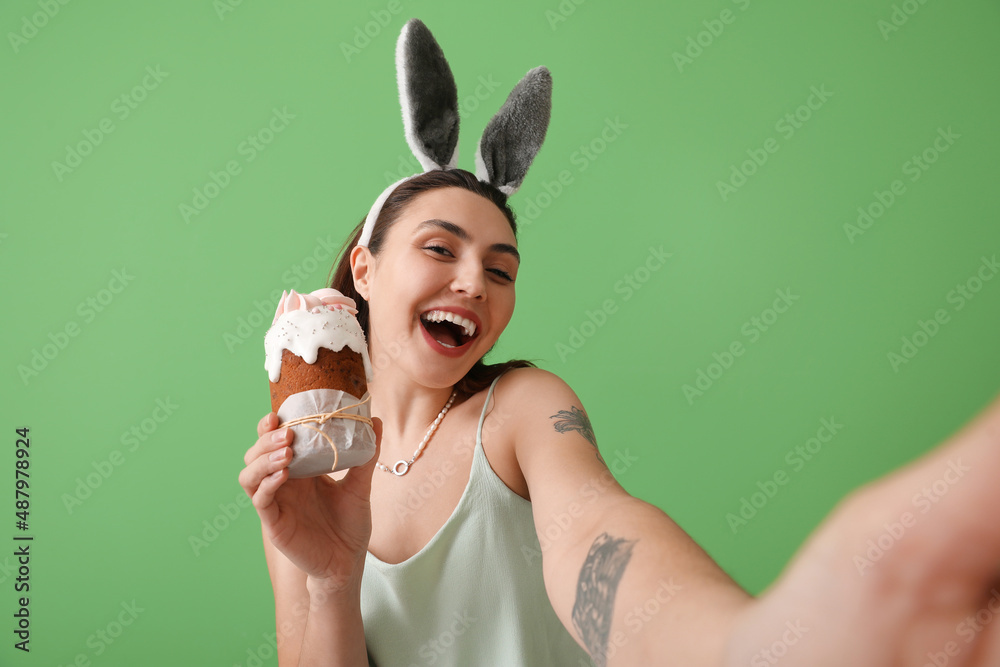 Young woman in bunny ears with tasty Easter cake on green background