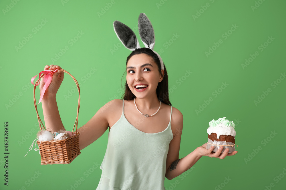 Young woman with tasty Easter cake and eggs in basket on green background