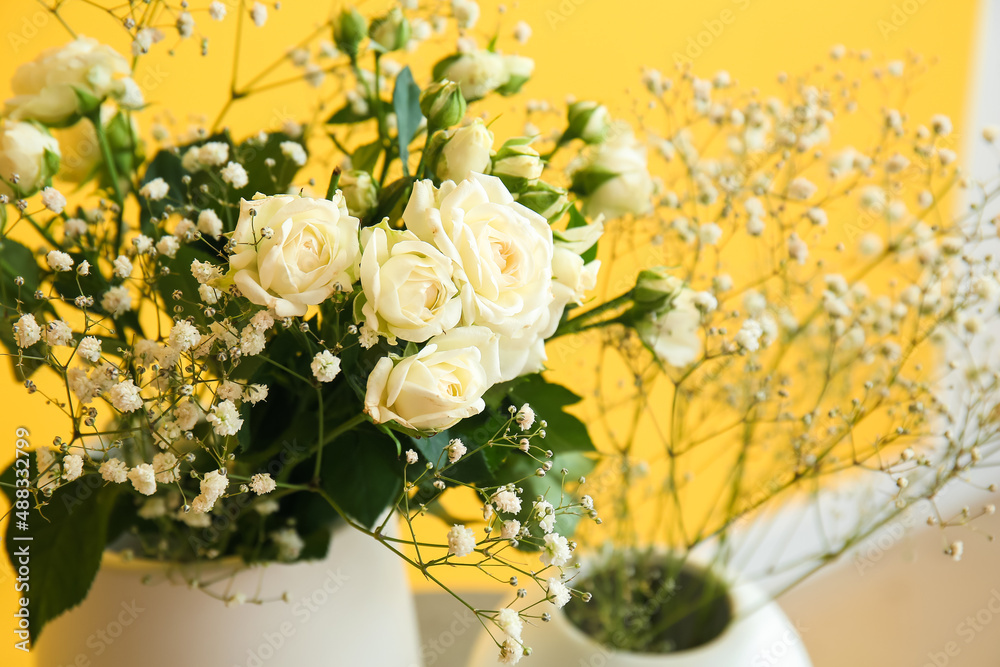 Vases with roses and gypsophila flowers near yellow wall, closeup