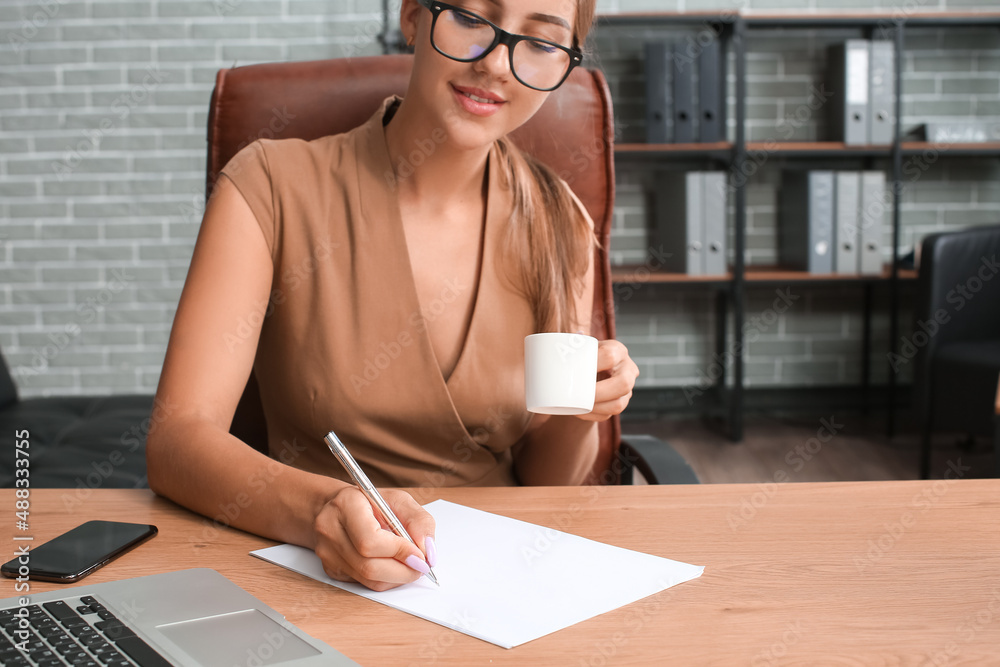 Young businesswoman with cup of coffee writing on paper sheet in office