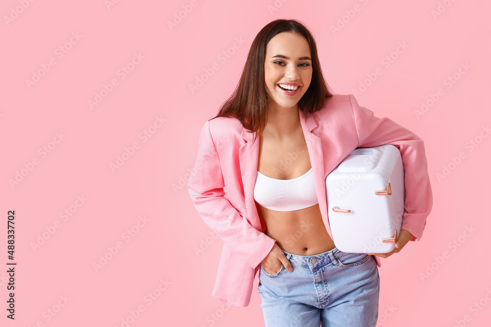 Beautiful young woman with small cosmetic refrigerator on color background