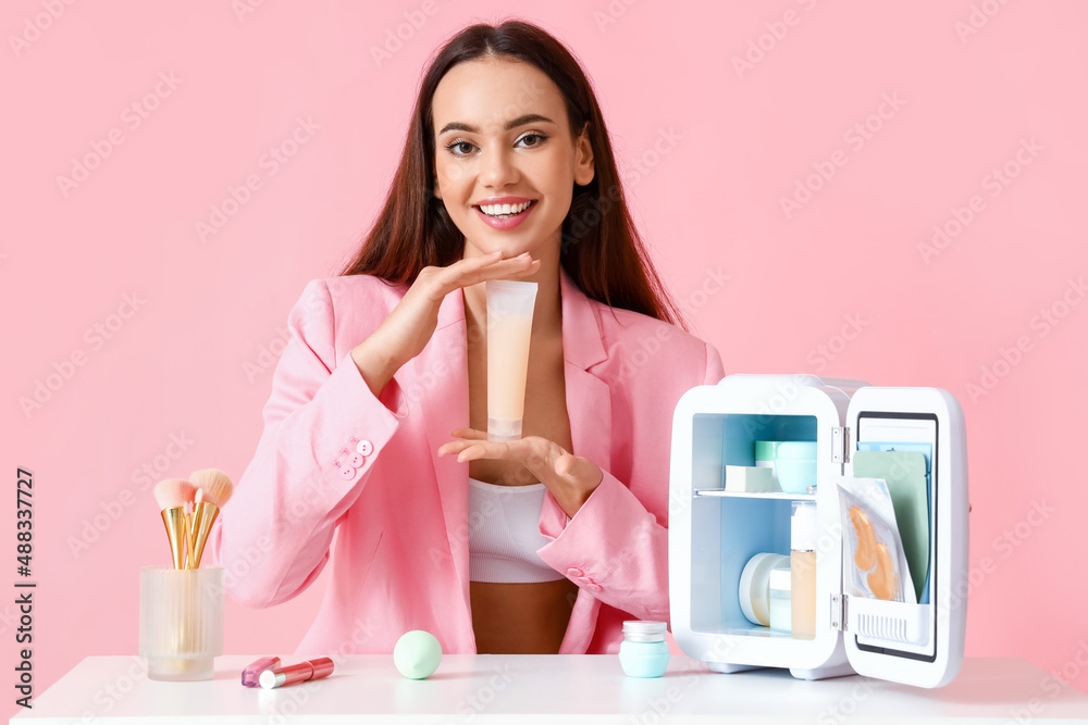 Beautiful young woman with small refrigerator and cosmetic products sitting at table on color backgr