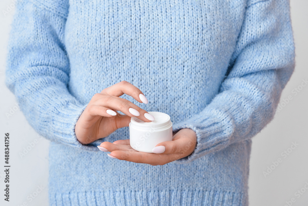 Woman in warm sweater holding jar with cream on light background, closeup