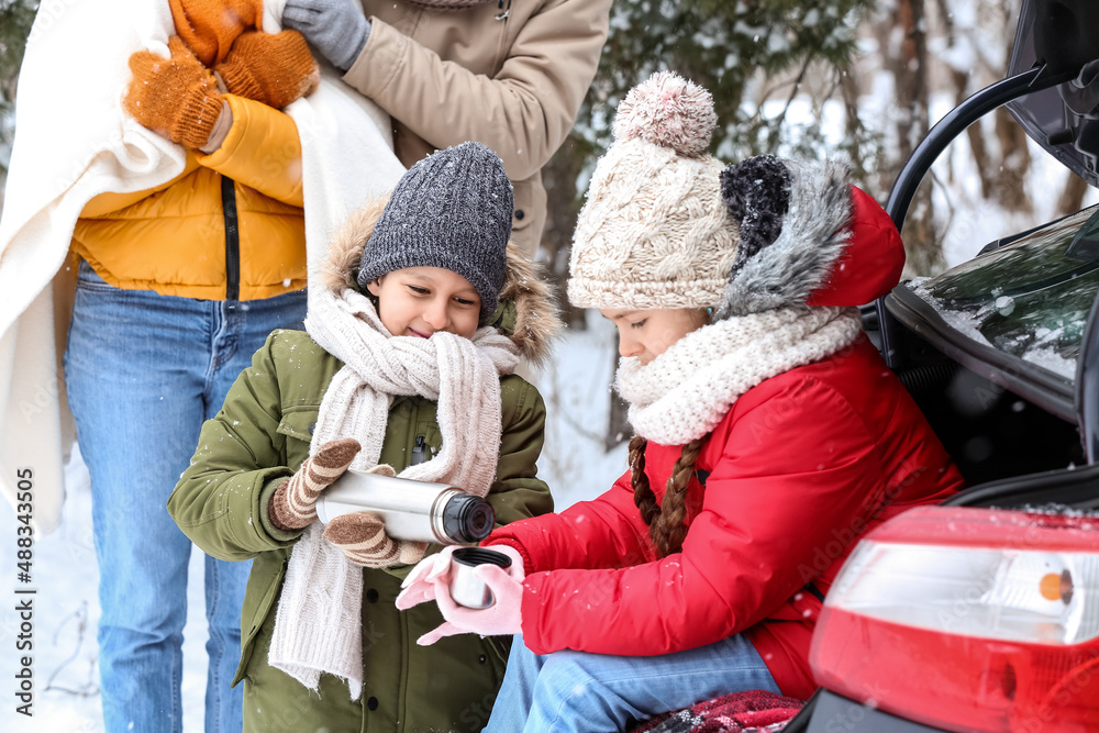 Happy family with thermos drinking hot tea near car in forest on snowy winter day