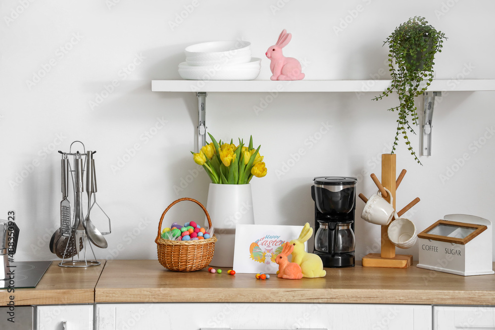 Vase with tulips, basket of Easter eggs, decor and different kitchen utensils on counter near light 