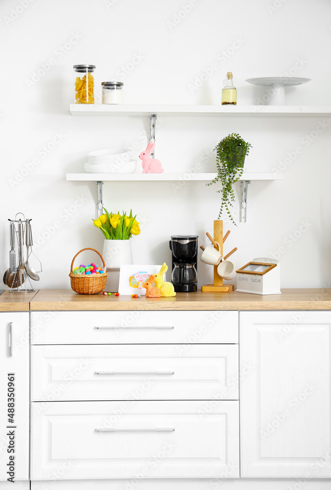 Basket with Easter eggs, rabbits, greeting card and vase with tulips on counter near light wall in k