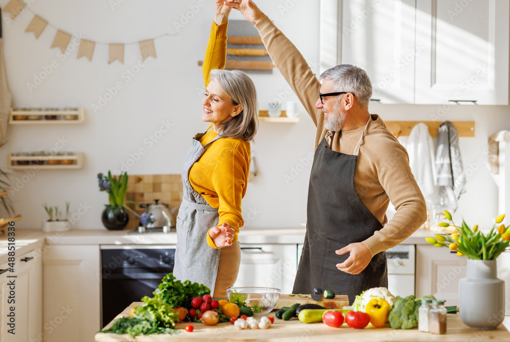 Happy senior elderly couple husband and wife embracing and dancing while cooking together in kitchen