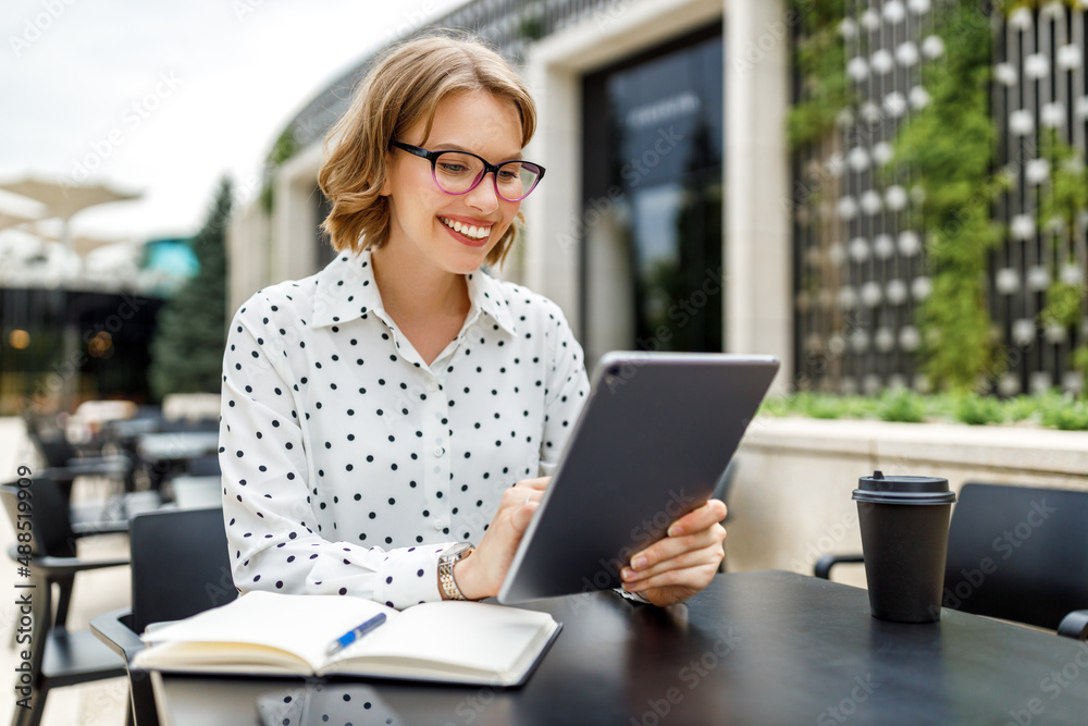 Lovely student girl in glasses sitting with digital tablet  and cup of takeaway coffee outside on te