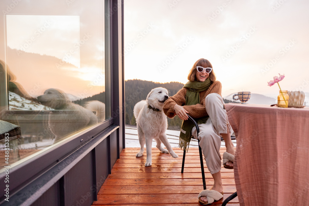 Woman playing with her dog while resting on terrace of tiny house in the mountains, enjoying beautif