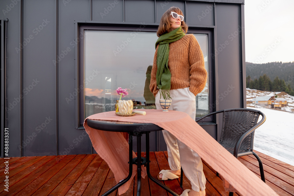 Young stylish woman standing on terrace of tiny house in the mountains, enjoying beautiful landscape