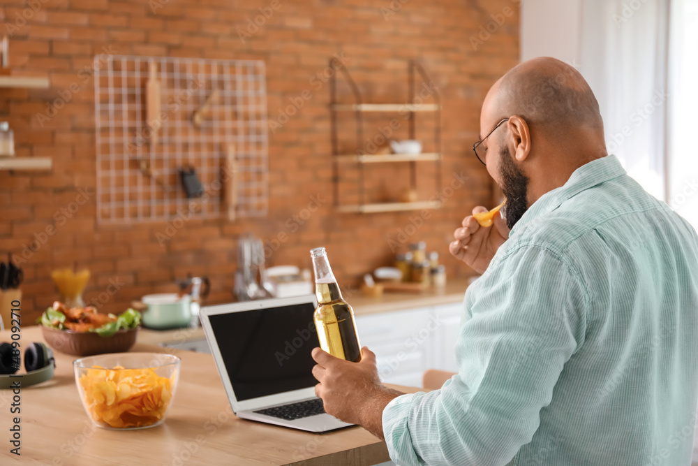 Bald man with laptop drinking beer and eating chips in kitchen