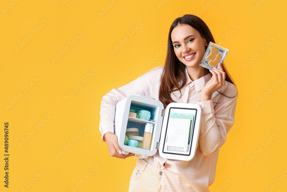 Beautiful young woman with small refrigerator and cosmetic products on color background