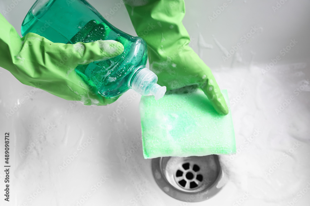 Woman in rubber gloves pouring detergent onto cleaning sponge near sink, closeup