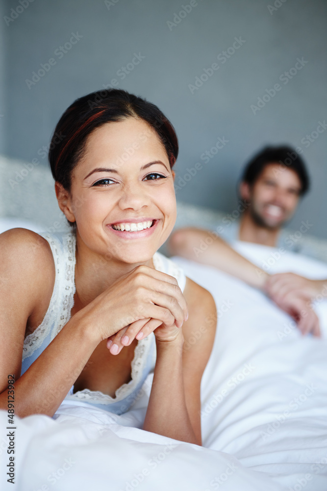 I love lying in. Portrait of a smiling woman lying on her bed with her boyfriend in the background.