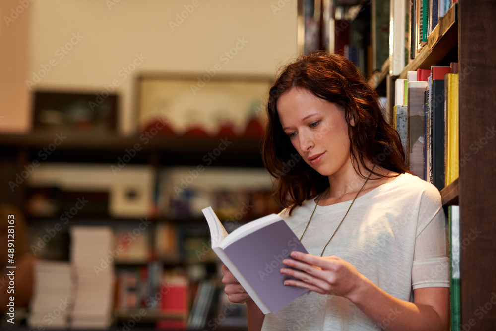 Absorbed in the plot. A young woman lost in the pages of a novel against a backdrop of full bookshel