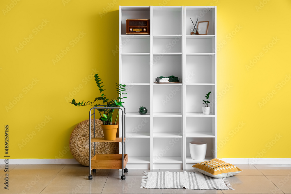 Big shelving unit with decor, houseplant and pouf near yellow wall