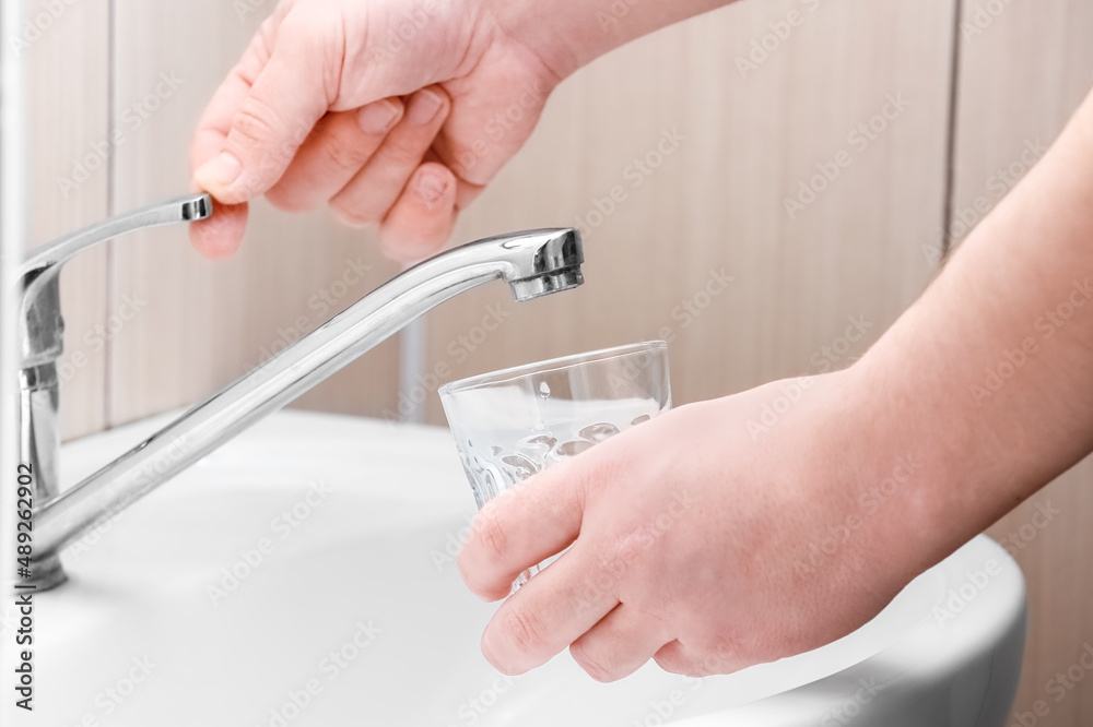 Man with glass turning on tap in bathroom, closeup