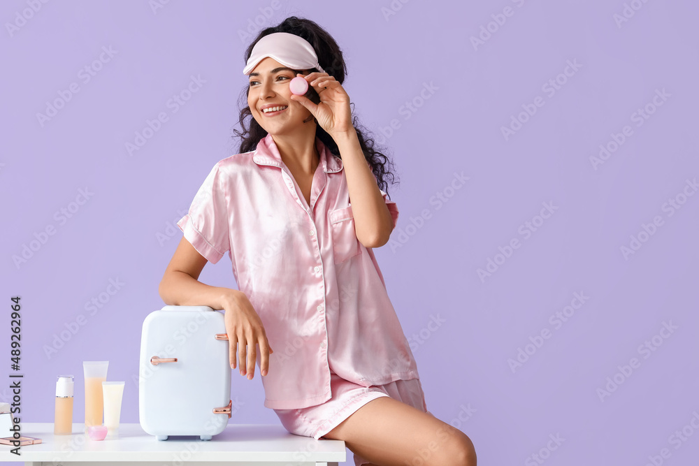 Young woman holding jar with cosmetic product near table with refrigerator on color background