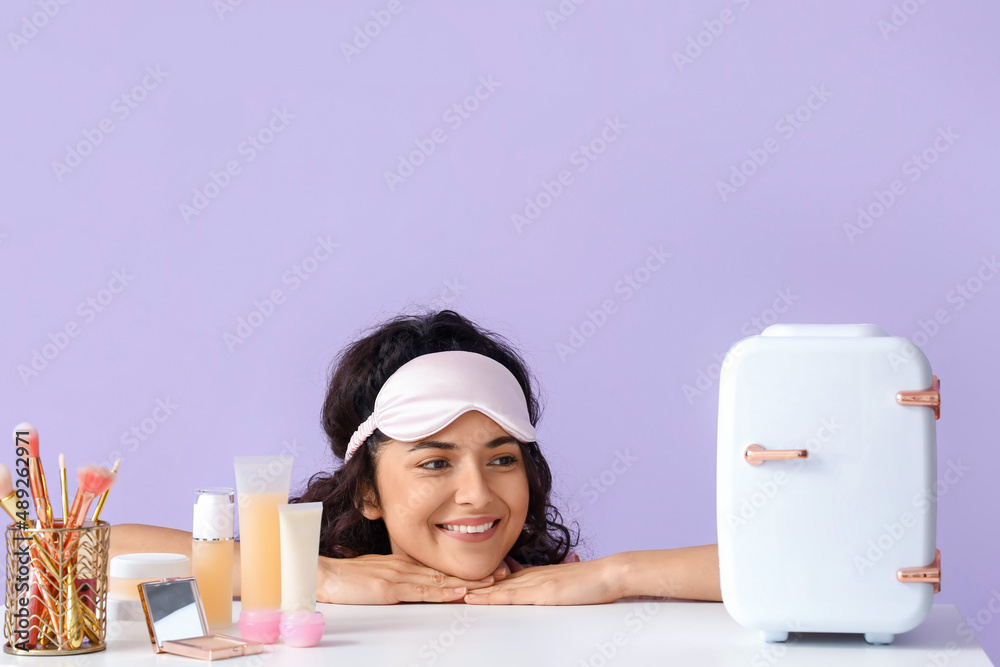 Young smiling woman with sleep mask near table looking at cosmetic refrigerator on color background