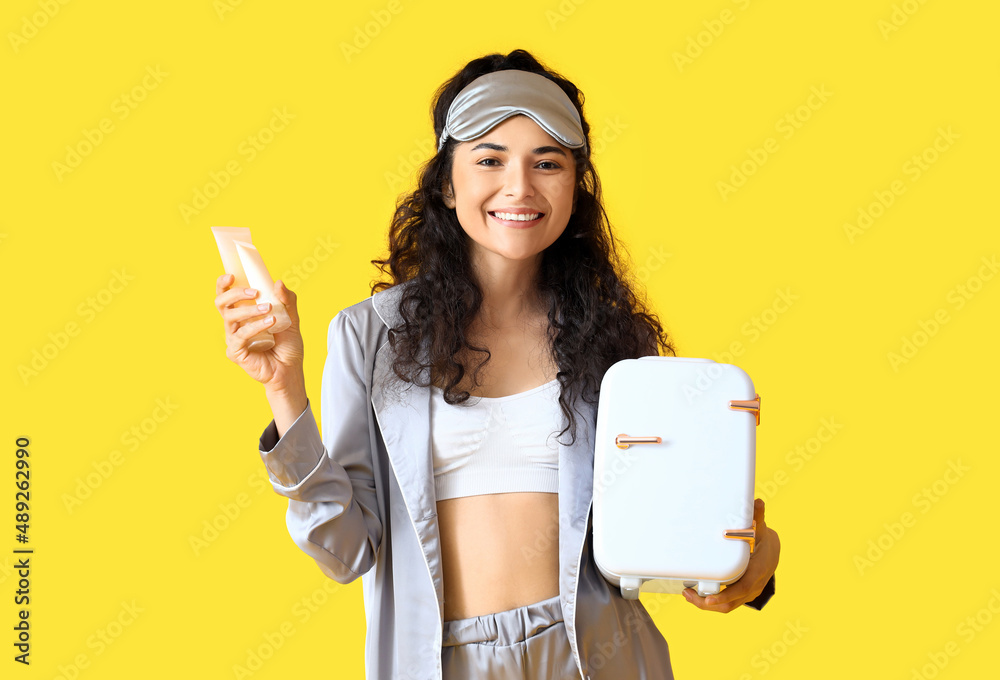 Young smiling woman in pajamas and sleep mask holding bottles with cosmetic products and refrigerato