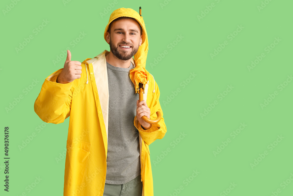 Young man in stylish raincoat and with umbrella showing thumb-up gesture on color background