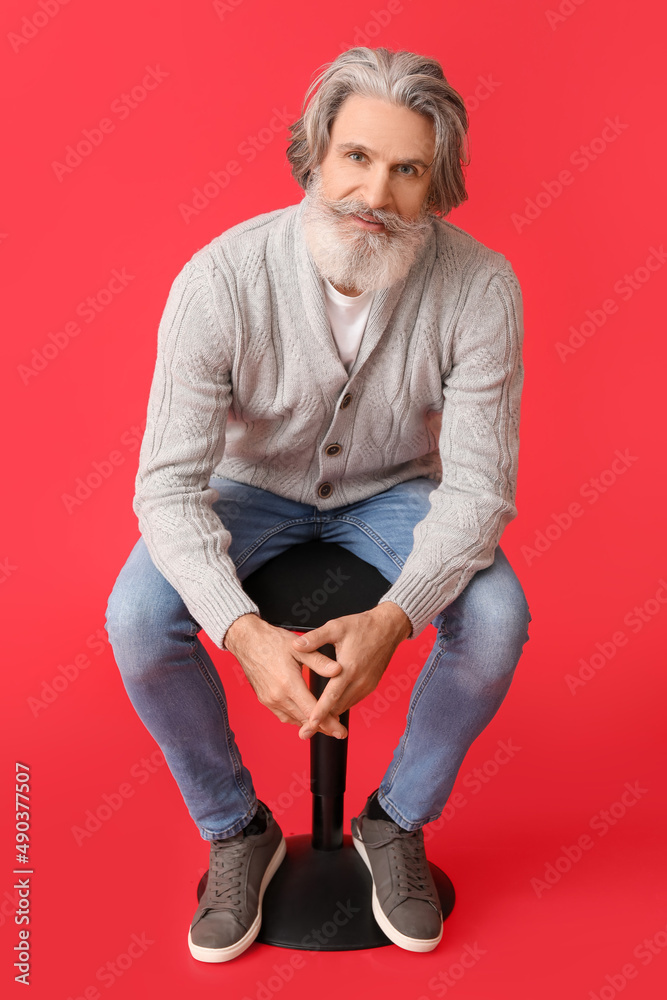 Handsome senior man in knitted sweater sitting on chair against red background