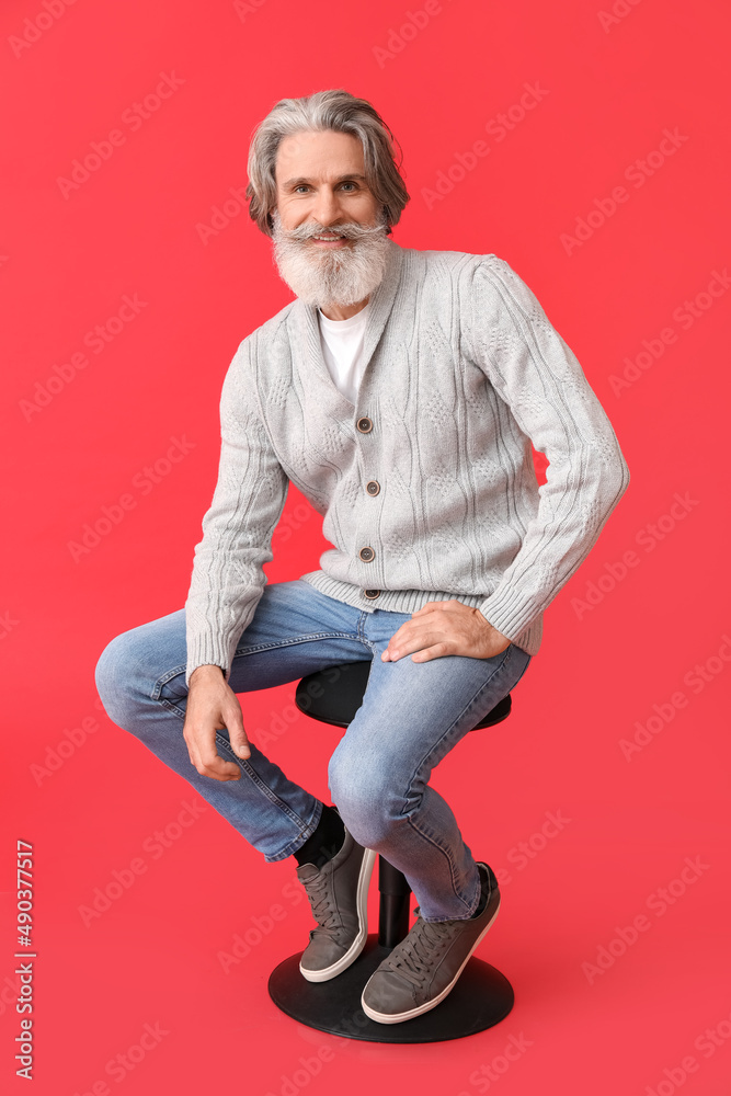 Handsome senior man in knitted sweater sitting on chair against red background