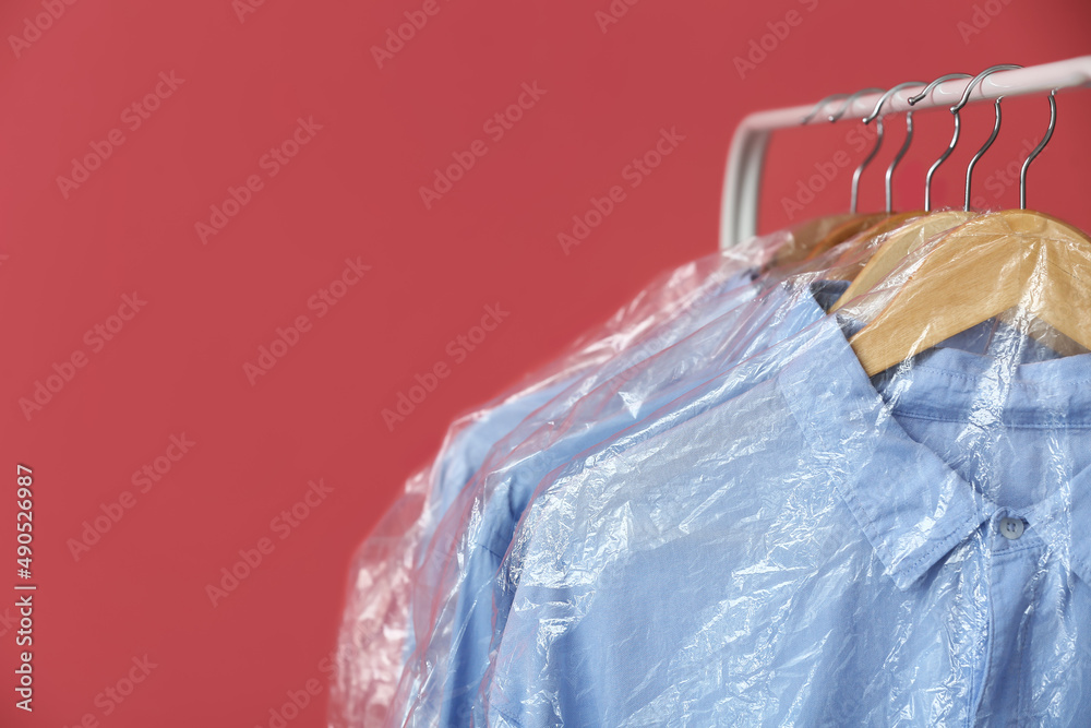 Rack with clean blue shirts in plastic bags on pink background, closeup