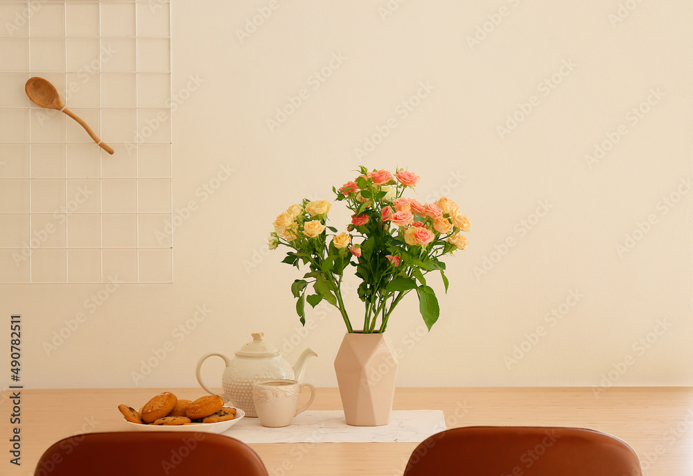 Vase with beautiful roses, teapot and cookies on dining table in kitchen