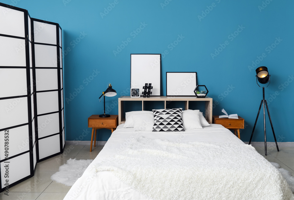 Comfortable bed, lamps, folding screen and blank posters on shelf near blue wall