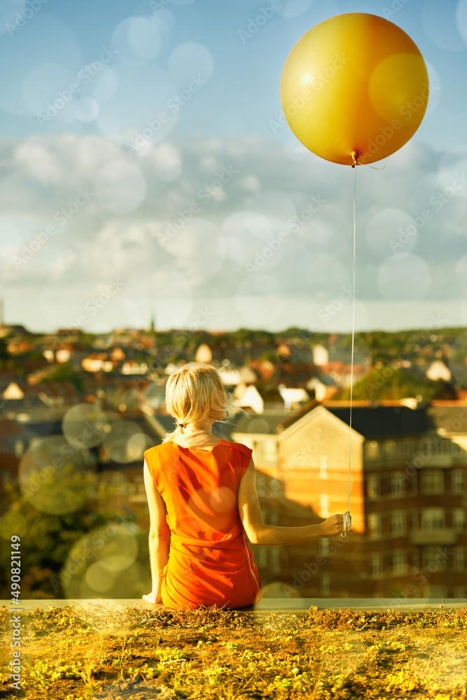 Reliving her childhood. Rear view of a young woman sitting with a balloon and a city in the backgrou