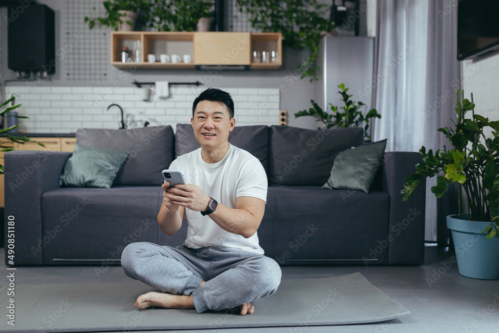 Male asian athlete at home using phone sitting on the floor during morning exercise, athlete in livi