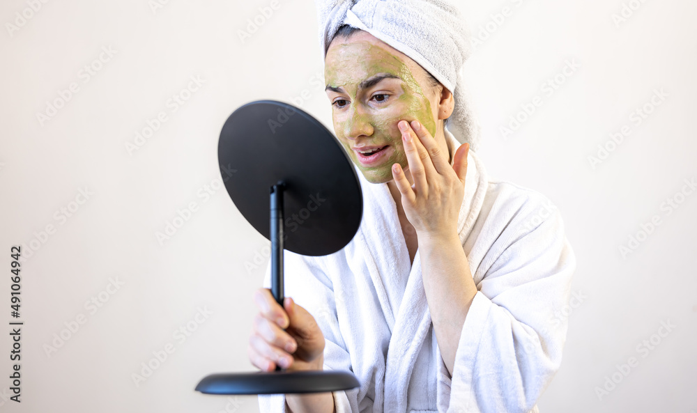 Young woman with a green mask on her face in a bathrobe in front of a mirror.