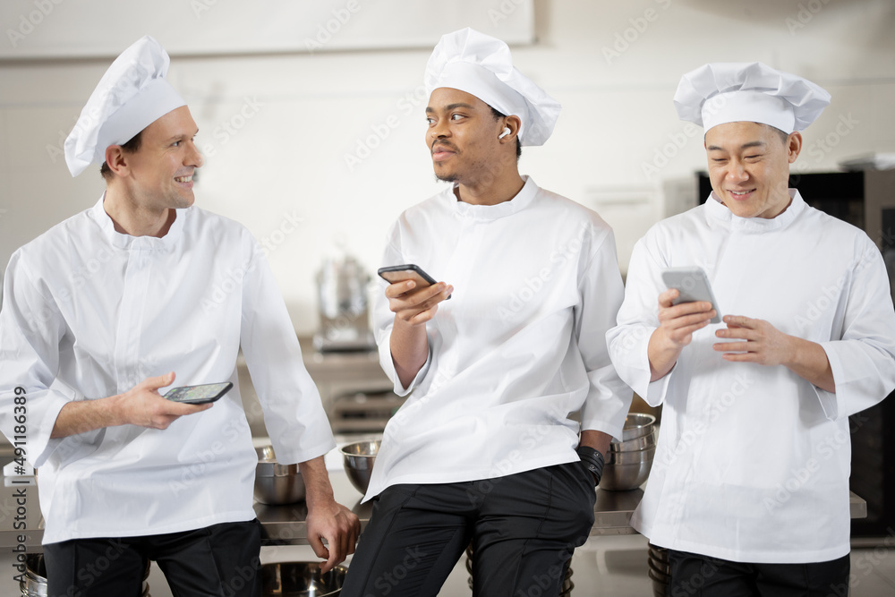 Three multiracial chef cooks talk while standing with smart phones during a break at the restaurant 