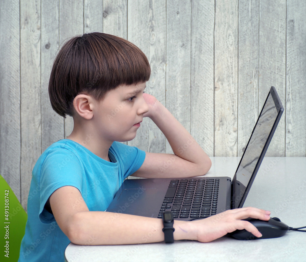 A nine-year-old caucasian boy in casual wear is sitting at a table and using a laptop