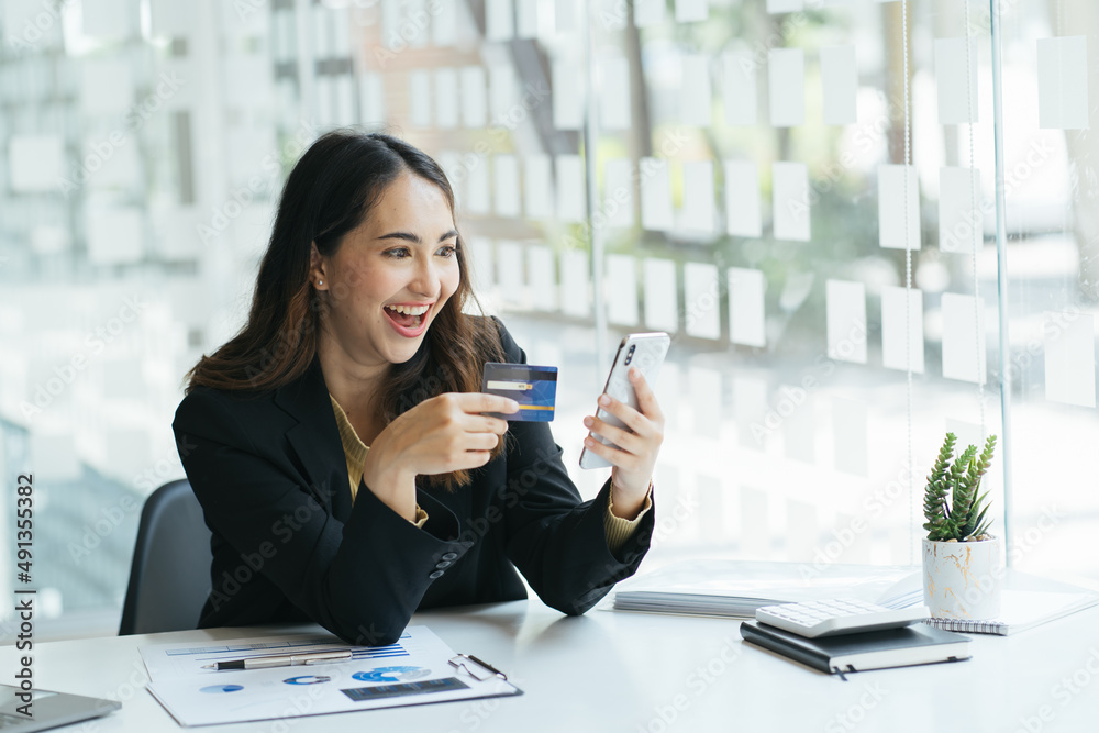 年轻漂亮的亚洲女人在办公室用智能手机和信用卡进行网上购物并复印