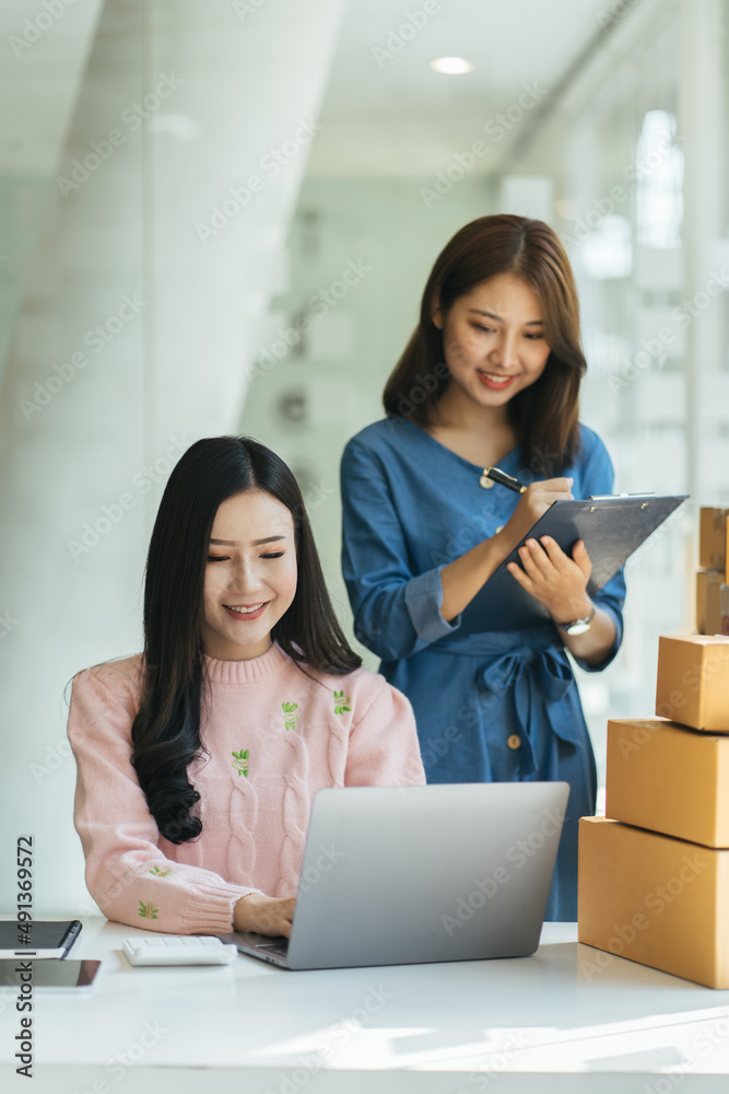 Two Asian women are looking at their laptop computers to check orders, with cardboard boxes and shop