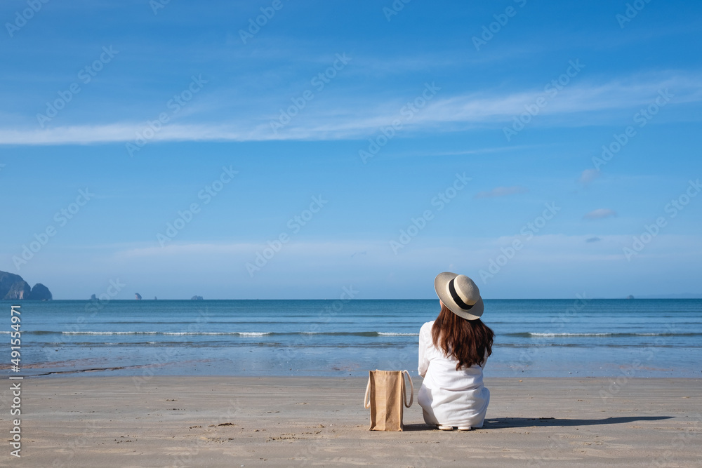 Rear view image of a woman with hat and bag sitting on the beach with blue sky background