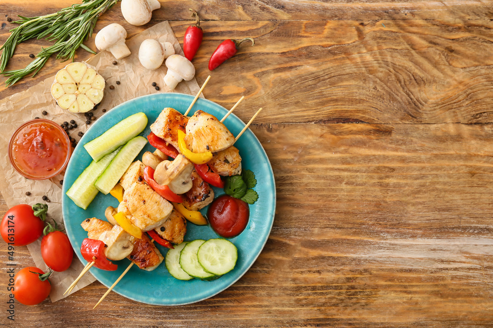 Plate of grilled chicken skewers with vegetables and sauce on wooden background