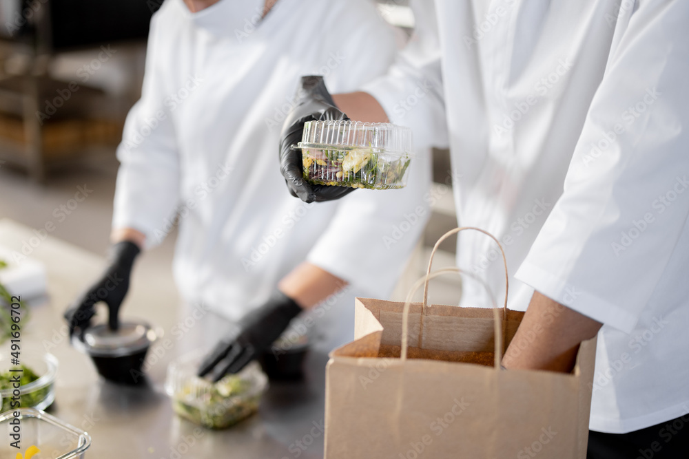 Cooks in protective gloves prepare take away food and packing them into paper bag for delivery, clos