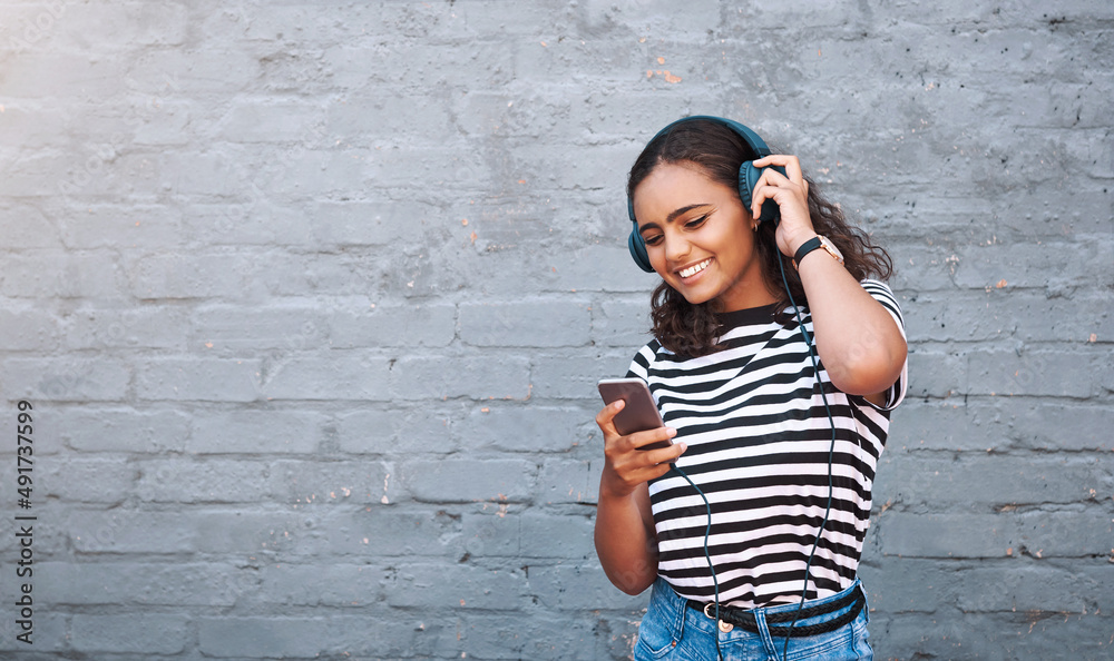 Getting her weekend playlist together. Shot of a young woman using a cellphone while wearing headpho