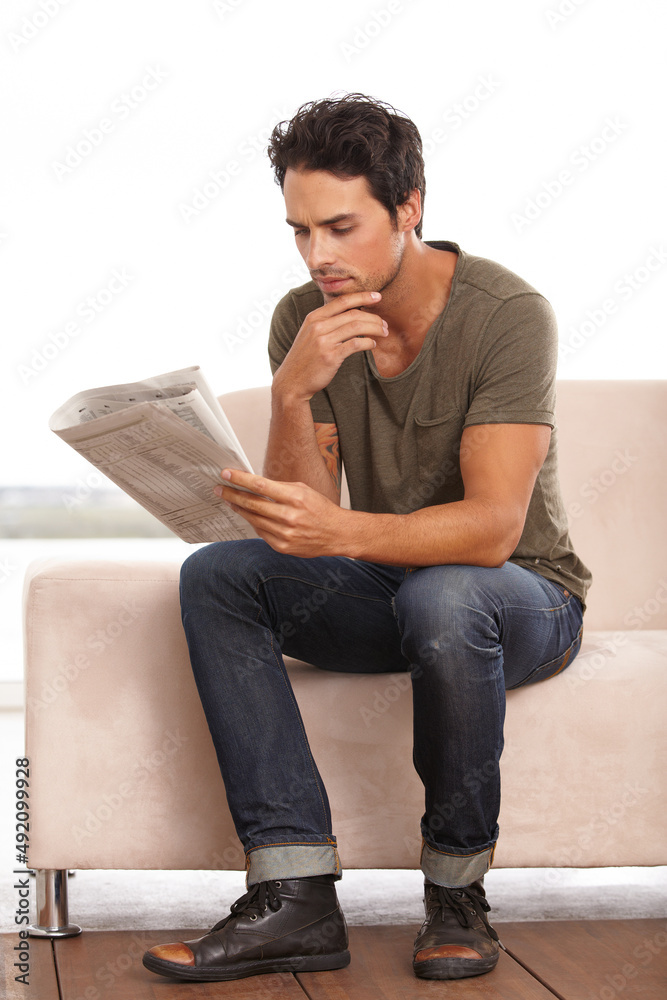 This is interesting. A handsome young man sitting on the couch at home reading a newspaper.