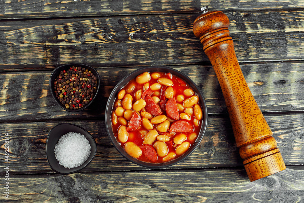 Beans baked with sausages. Beans with fried sausages in tomato sauce. Food in a black bowl on a brow