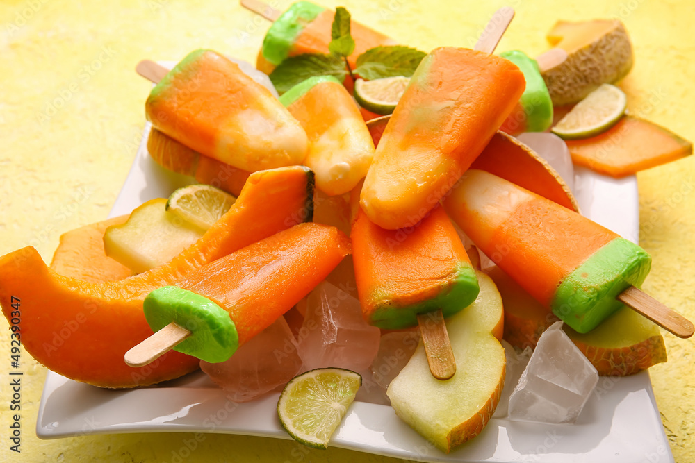 Plate with tasty popsicles and melon pieces on yellow background