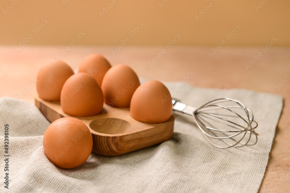 Holder with fresh chicken eggs and whisk on beige background