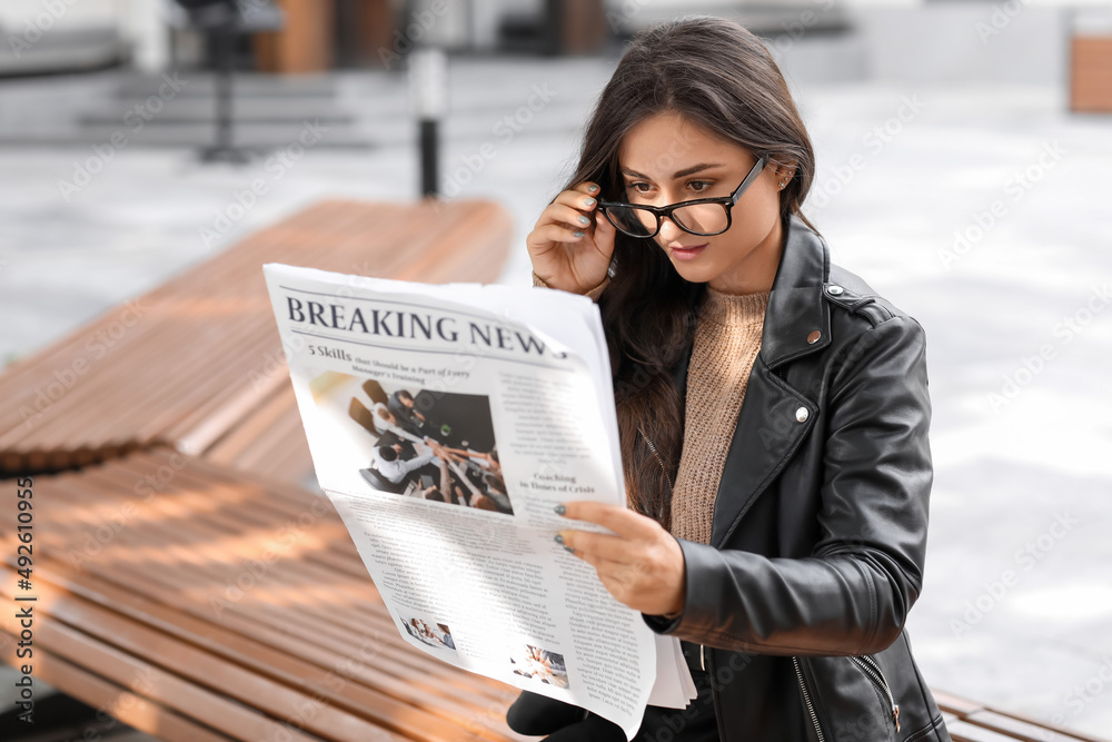 Young woman in eyeglasses reading newspaper outdoors