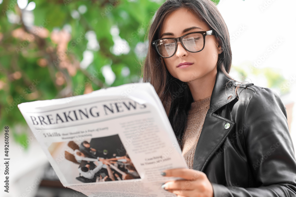 Young woman in eyeglasses reading newspaper outdoors