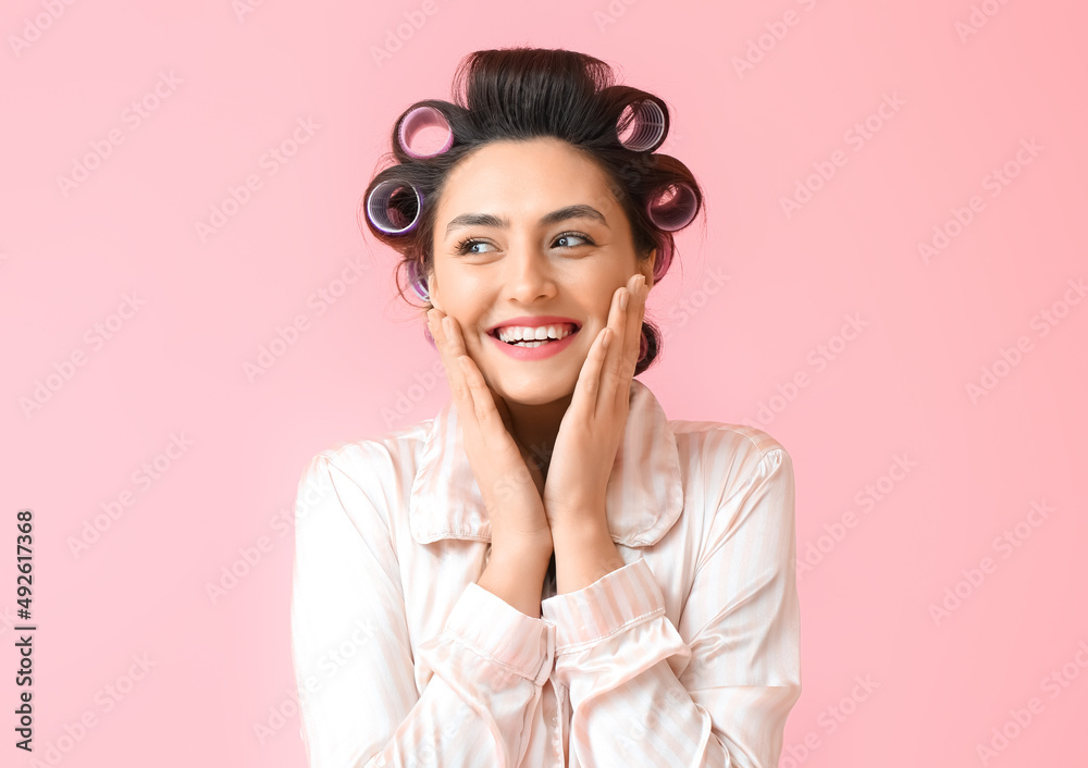 Beautiful young woman in pajamas and hair curlers on pink background