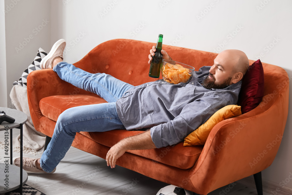 Funny lazy man drinking beer and eating chips while watching TV at home